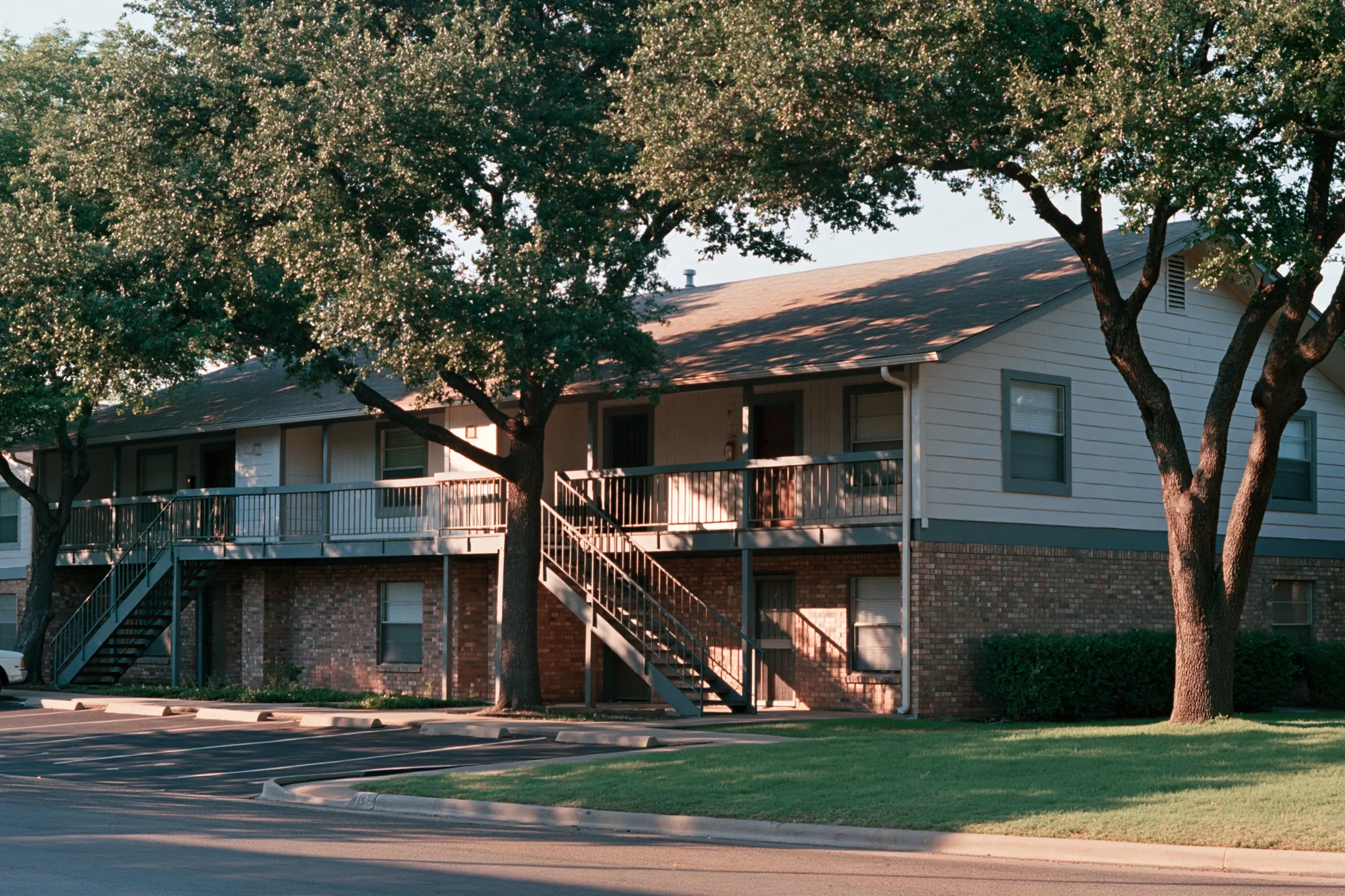 Two-story 1980s garden apartment building with exterior walkways, wood siding and brick, live oaks, no people